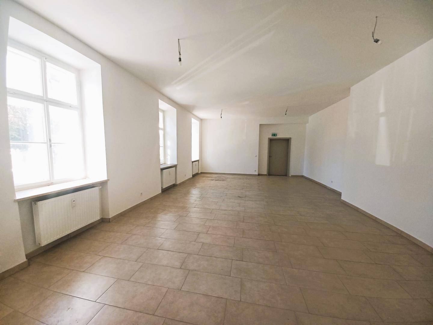 Geräumiger, leerer Raum mit großen Fenstern, Fliesenboden und Heizkörpern unter den Fenstern.