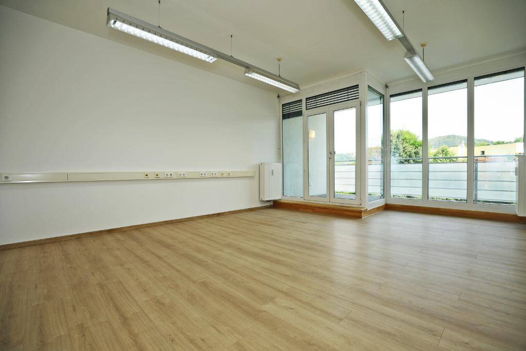 Heller Büroraum mit Laminatboden, weißen Wänden und breiter Fensterfront, die viel Licht spendet.