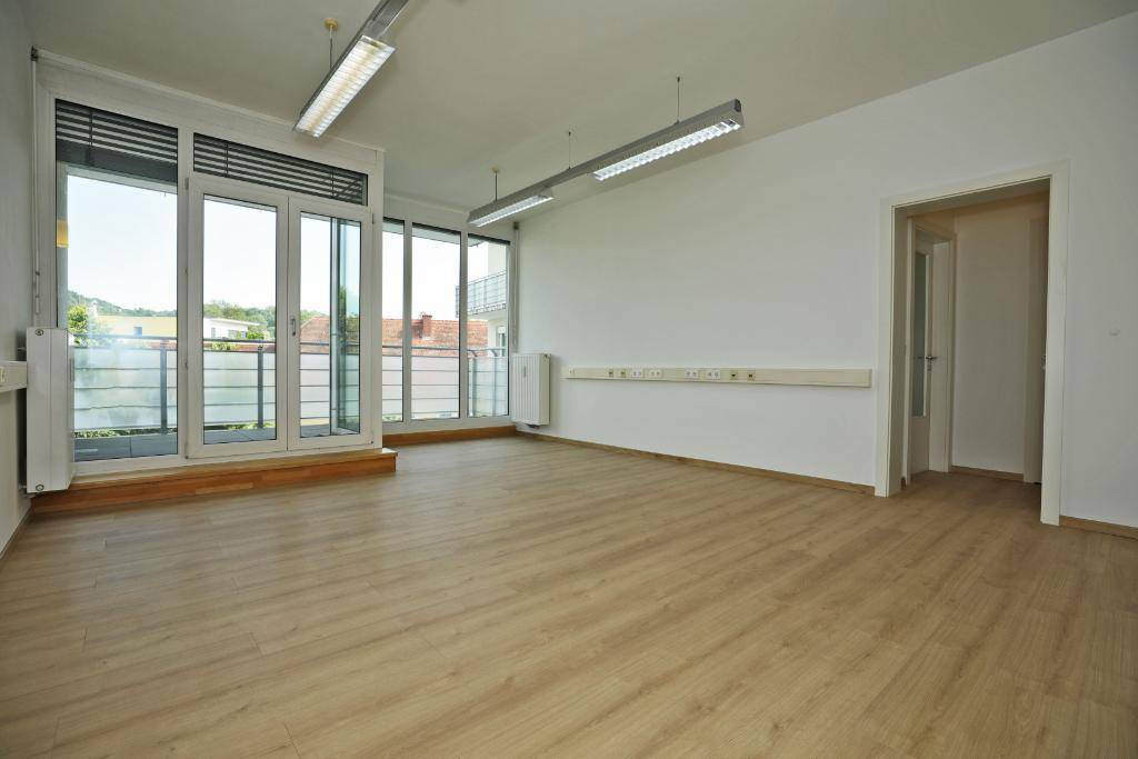 Geräumiger Büroraum mit hellem Laminatboden, weißen Wänden und großen Fenstern zum Balkon.