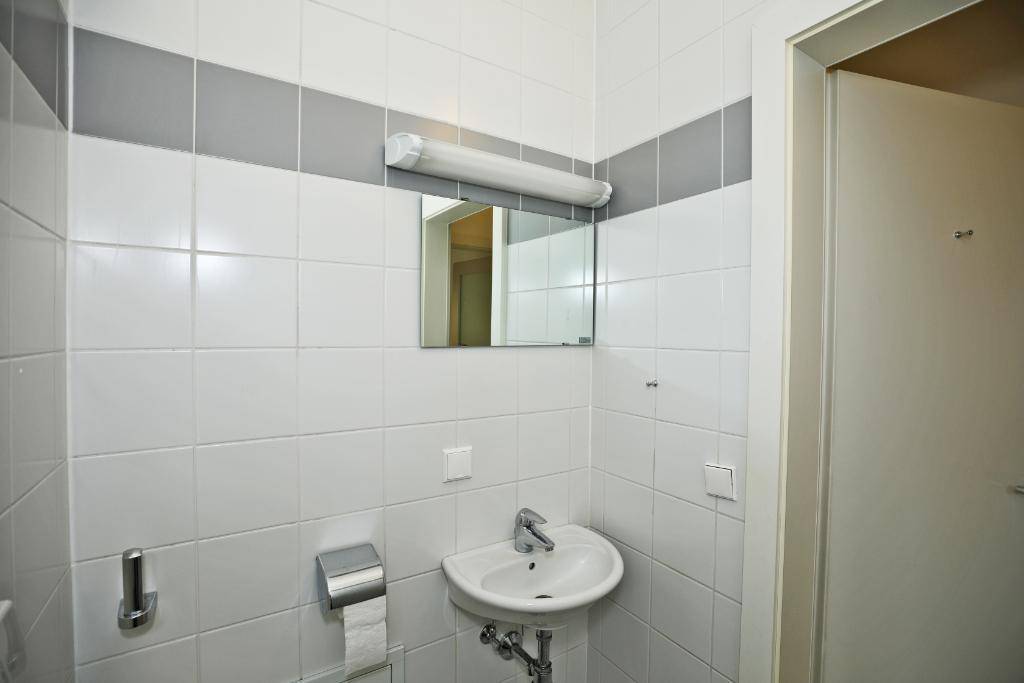 Sauber gefliestes WC mit Waschbecken und Spiegel, in schlichtem Weiß und Grau gehalten.