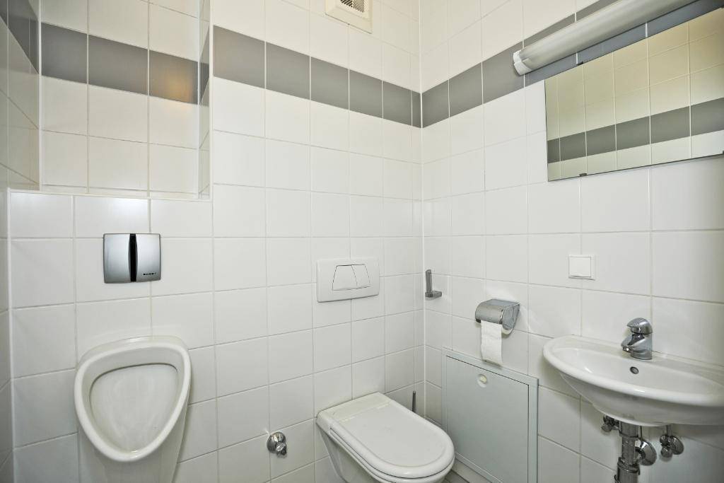 Modern gefliestes WC mit Urinal, Toilette und kleinem Waschbecken in Weiß und Grau.