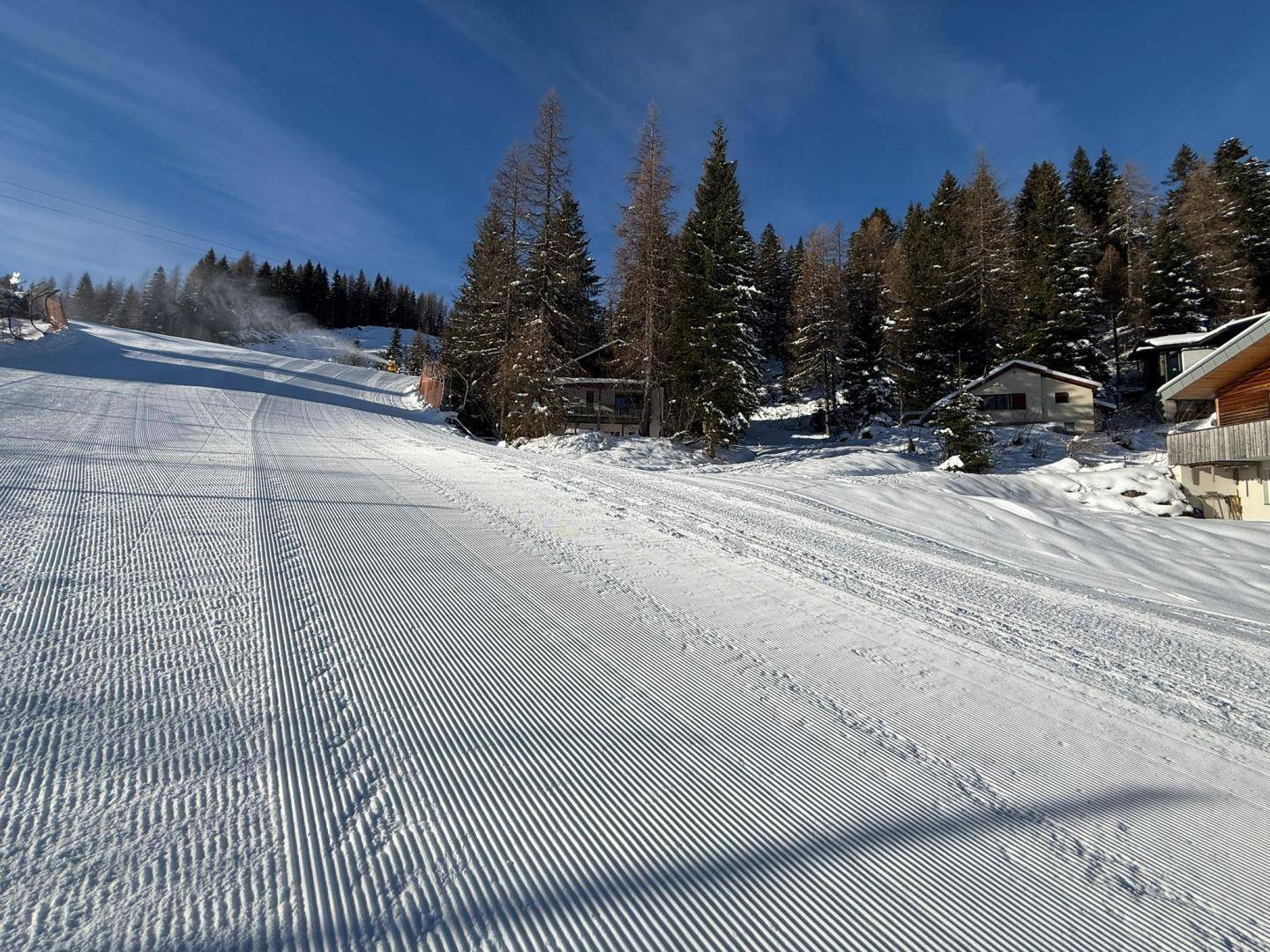 Eine frisch präparierte Skipiste führt durch eine verschneite Landschaft mit Häusern und Bäumen im Hintergrund.