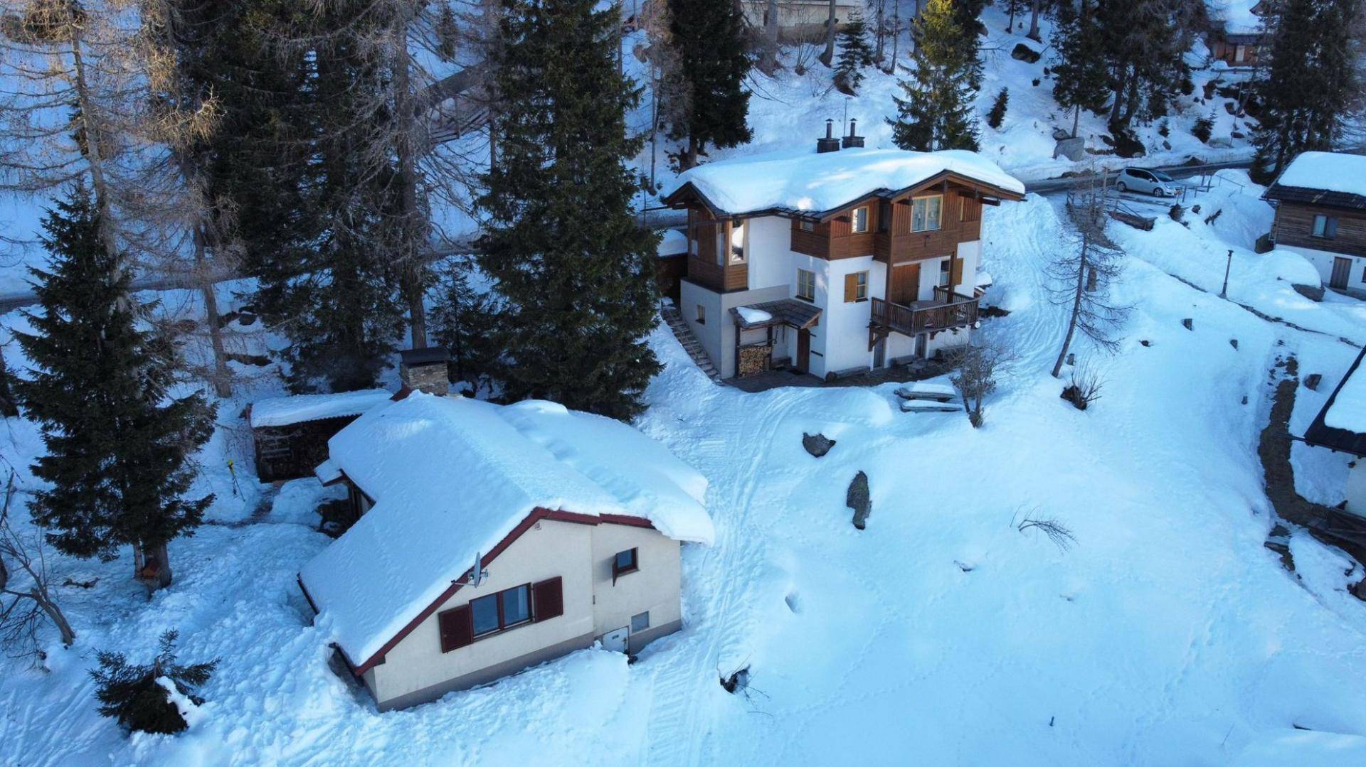 Luftaufnahme von zwei Häusern in einer tief verschneiten Berglandschaft, umgeben von Nadelbäumen.