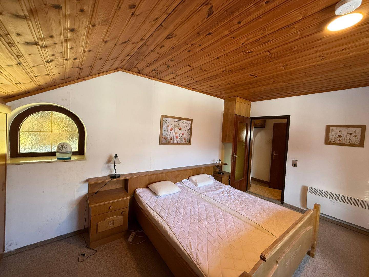 Helles Schlafzimmer mit Holzdecke, Doppelbett und einem Fenster mit dekorativem Bogen.