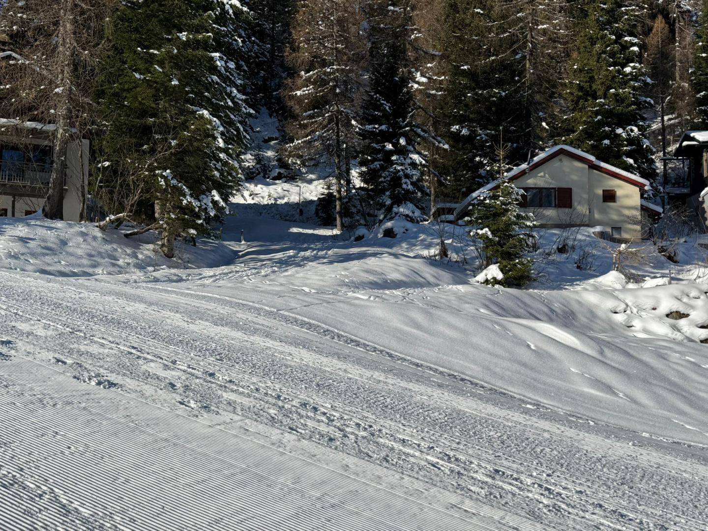Eine frisch präparierte Skipiste führt durch eine verschneite Landschaft mit Häusern und Bäumen im Hintergrund.