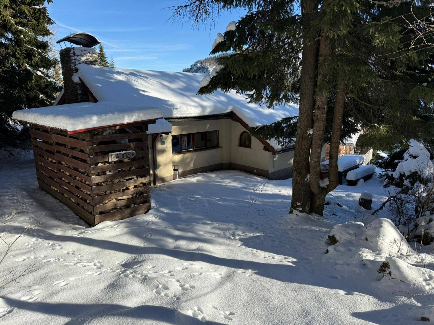 Verschneites Haus in alpiner Umgebung mit Holzlager und hohen Nadelbäumen unter blauem Himmel.