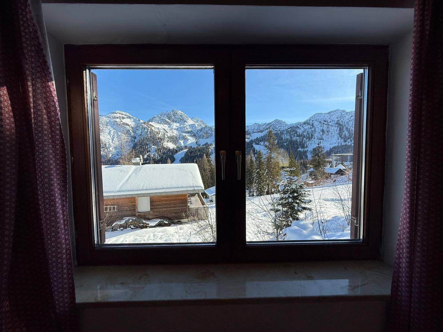 Fensterblick auf eine verschneite Berglandschaft mit sonnigem Himmel und einem weiteren Haus im Vordergrund.