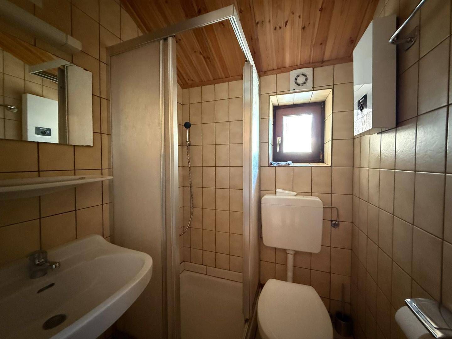 Kleines Badezimmer mit beigen Fliesen, Dusche, WC und einem Fenster für Tageslicht.