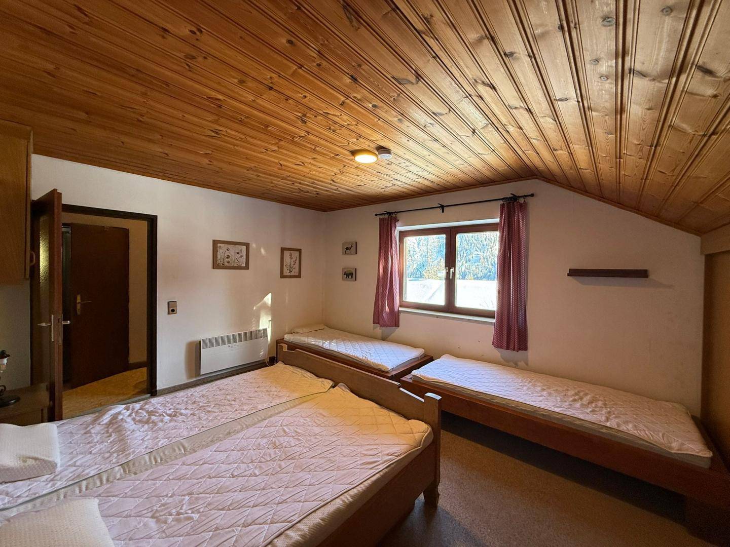 Schlafzimmer mit Holzdecke, drei Einzelbetten und einem Fenster mit Blick auf die verschneite Landschaft.
