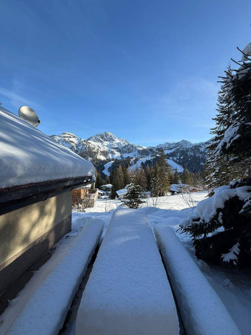Verschneiter Außensitzbereich mit Holztisch und Bänken, umgeben von Bäumen und einem beeindruckenden Bergblick.