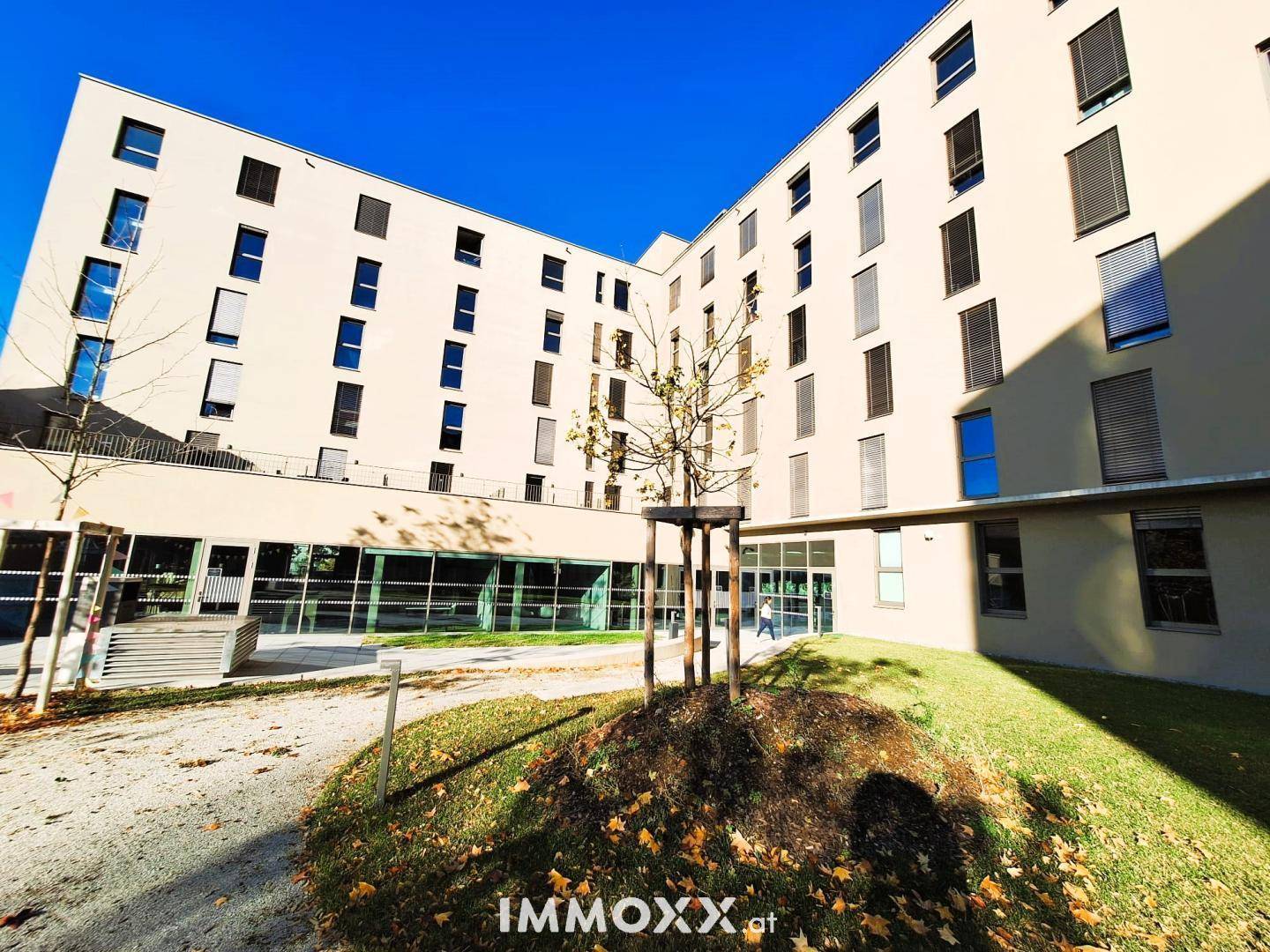 Außenansicht eines modernen Wohngebäudes mit begrüntem Innenhof und Bäumen unter blauem Himmel.