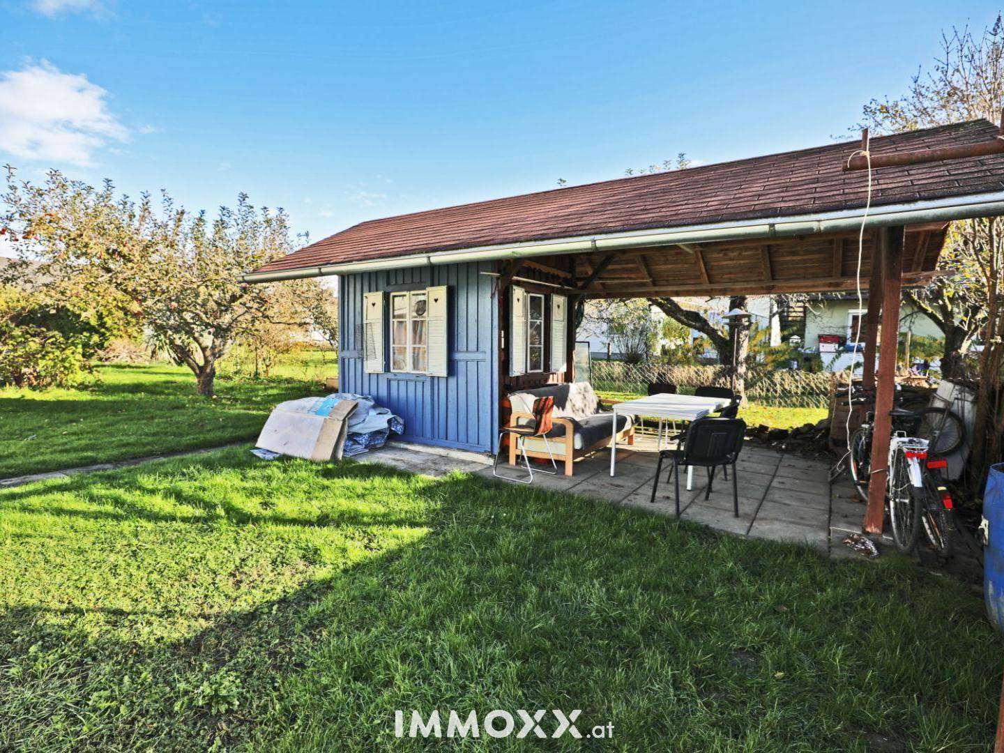 Überdachter Sitzbereich im Garten mit Holzhütte und Blick auf die grüne Umgebung.