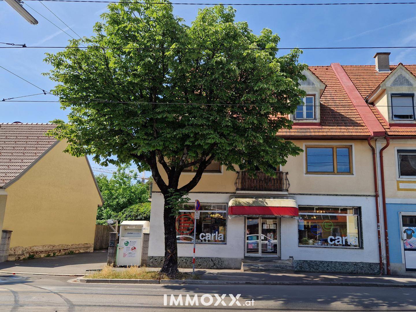 Fassade eines Gebäudes mit Geschäft im Erdgeschoss und Wohnungen darüber, umgeben von Grün.