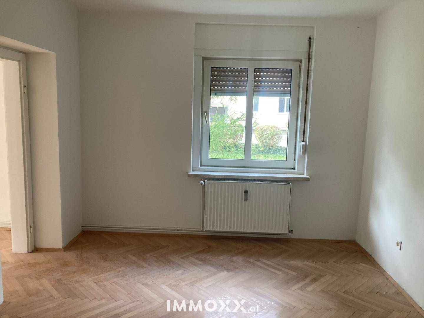Leerer Raum mit Fischgrätparkett, weiß gestrichenen Wänden und einem Fenster mit Heizkörper.