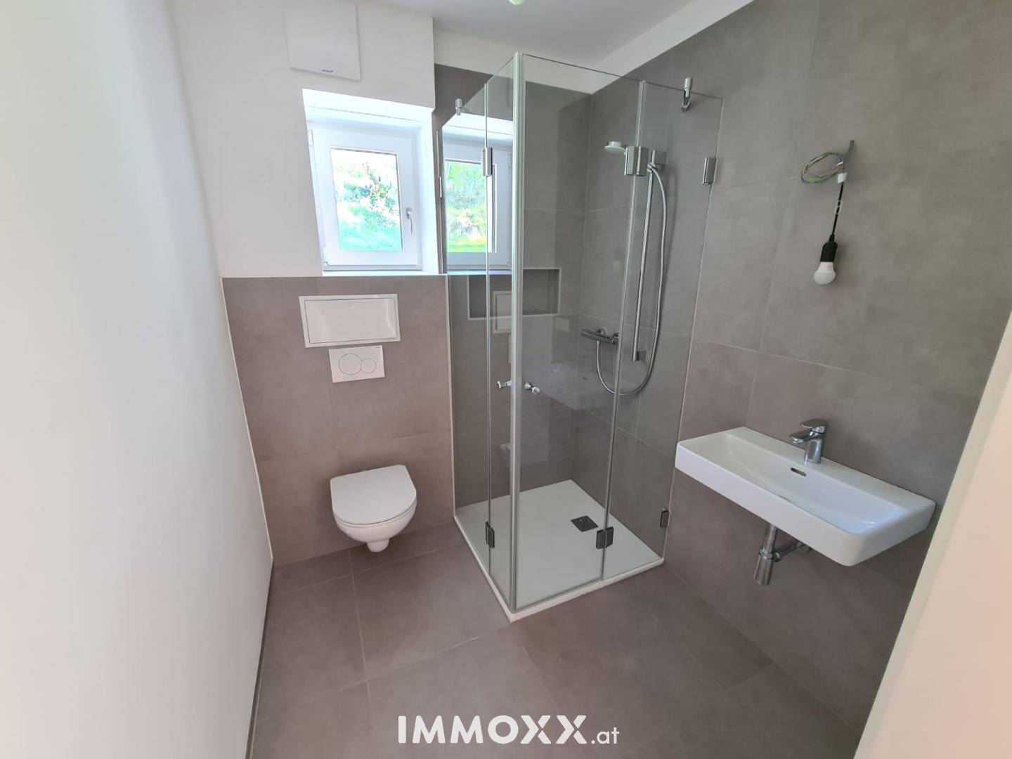 Modernes Badezimmer mit grauen Fliesen, ebenerdiger Dusche und Fenster für Tageslicht.