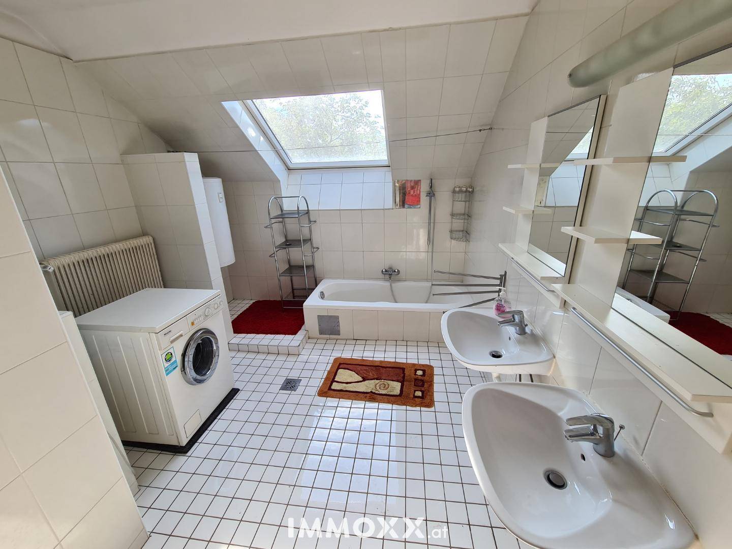 Helles Badezimmer mit weißen Fliesen, Badewanne, Doppelwaschbecken, Waschmaschine und Dachfenster.
