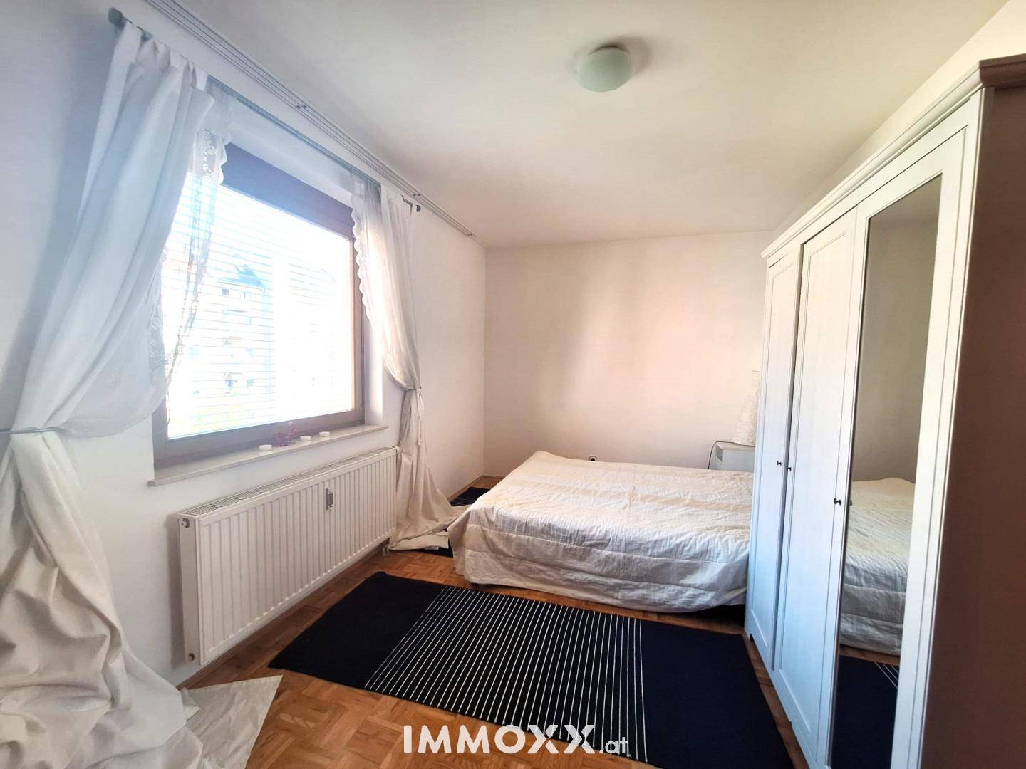 Schlafzimmer mit Parkettboden, einem Doppelbett und einem großen Fenster mit hellen Vorhängen.