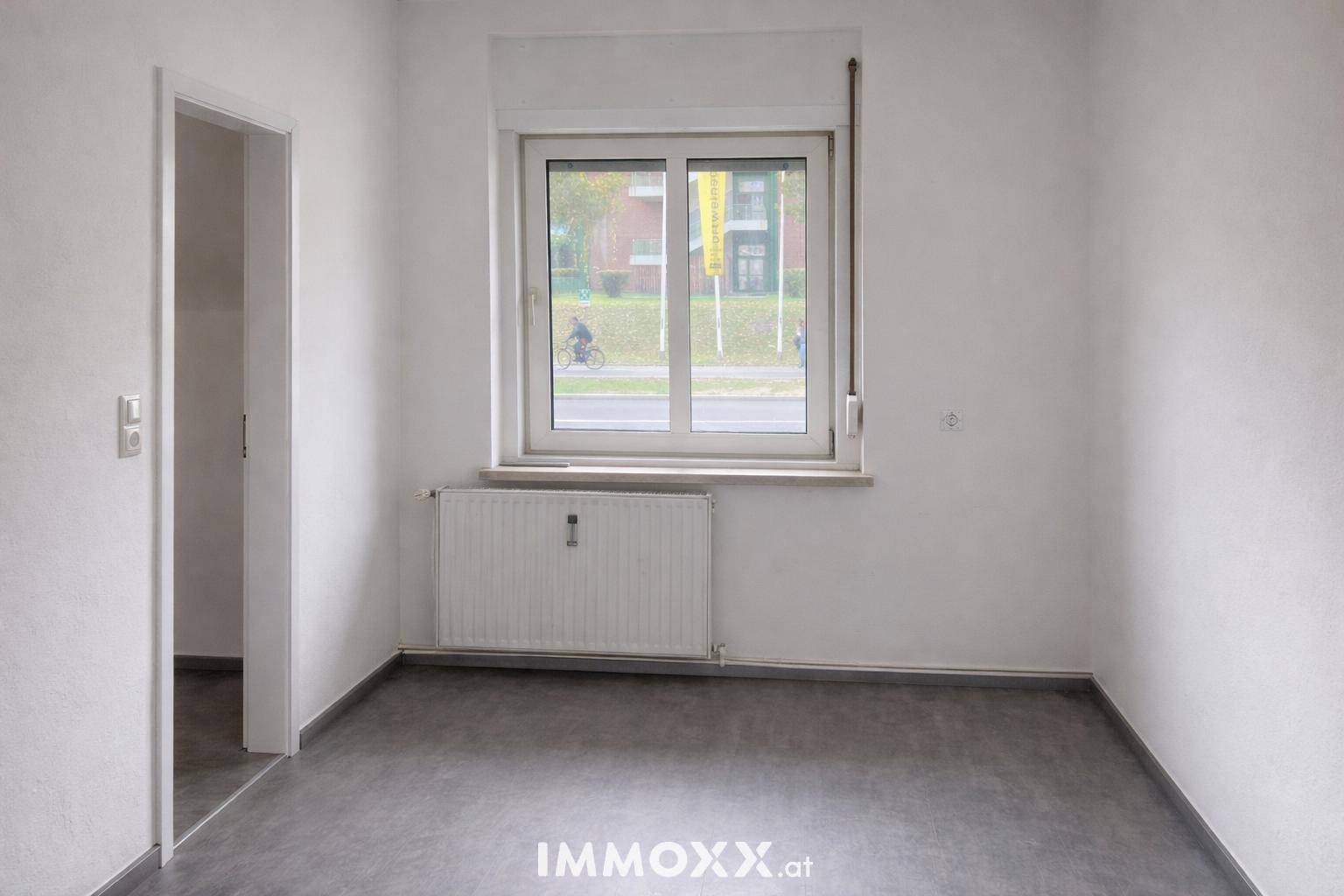 Leerer Raum mit grauem Boden, weiß gestrichenen Wänden und einem Fenster mit Heizkörper darunter.