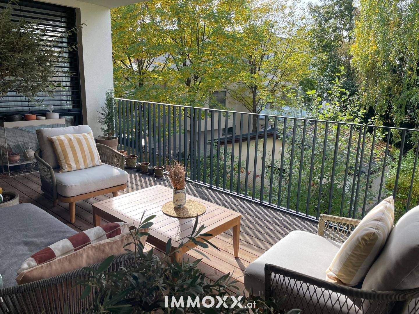 Sonniger Balkon mit Holzboden, bequemen Outdoor-Möbeln und Blick auf die umliegenden Bäume.