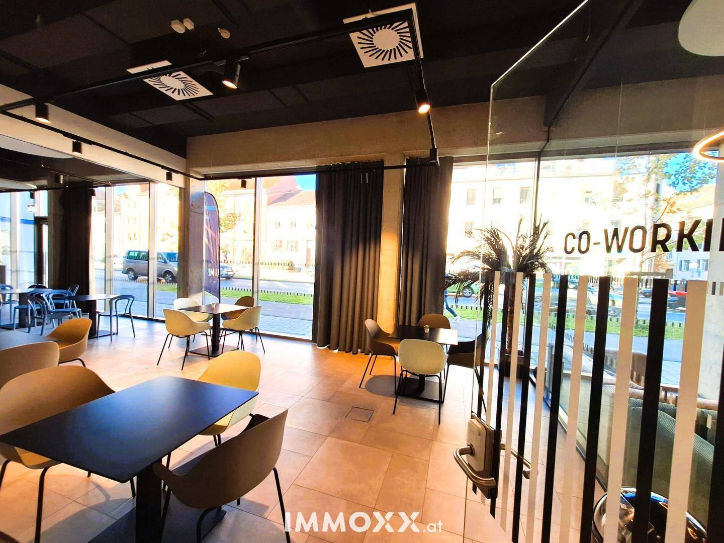 Moderner Co-Working-Bereich mit Tischen, Stühlen und großen Fenstern, die viel Licht hereinlassen.