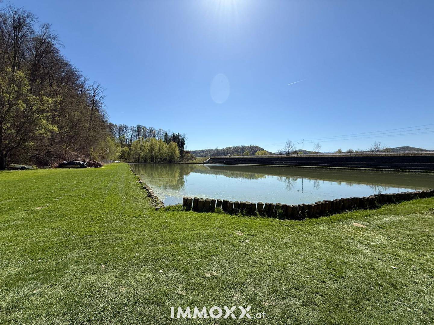 Panoramablick auf einen ruhigen Teich, umgeben von grünen Wiesen und einem Waldrand unter blauem Himmel.