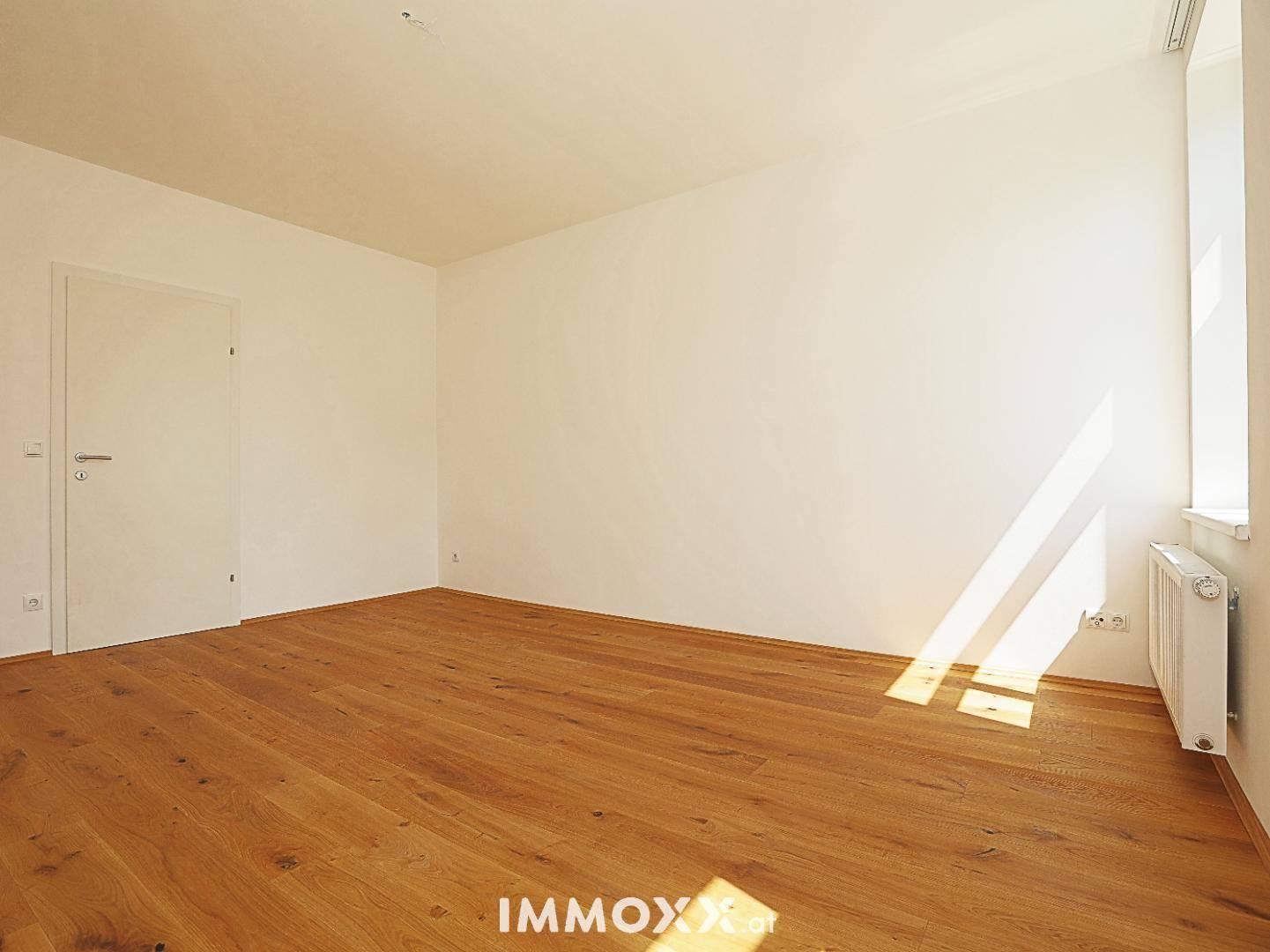 Geräumiger, heller Raum mit Holzboden und einem Fenster, das Sonnenlicht auf den Boden wirft.