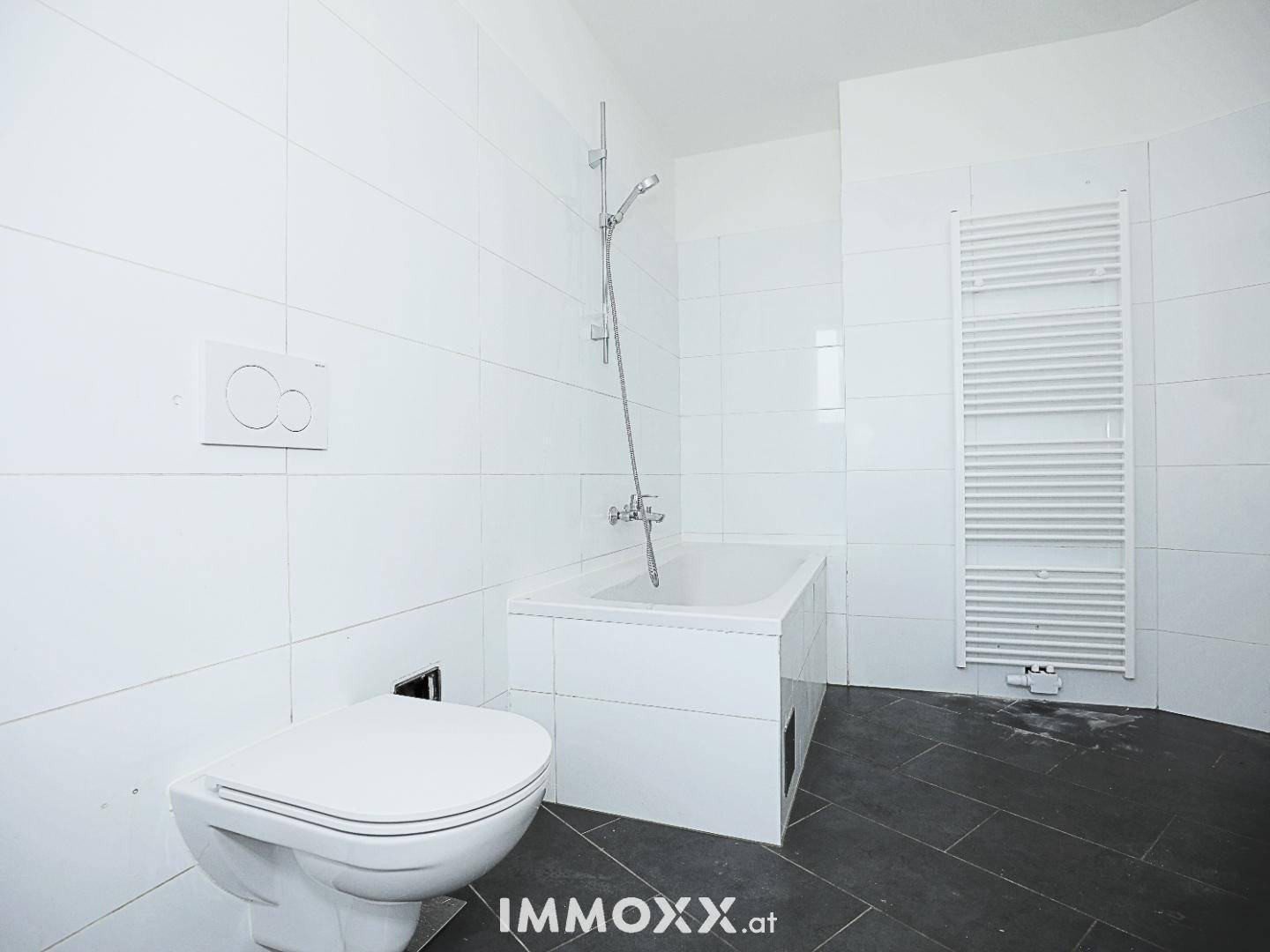 Badezimmer mit Toilette, Badewanne und Duscharmatur, umgeben von weißen Fliesen und dunklem Boden.