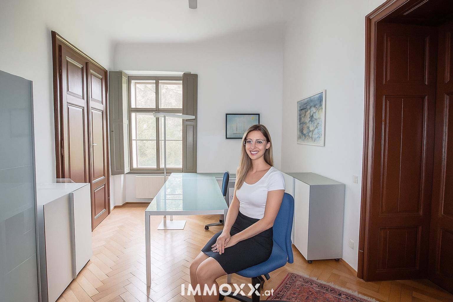 Helles Büro mit Fischgrätparkett, großen Fenstern und eleganten Holztüren, eine Frau sitzt am Schreibtisch.