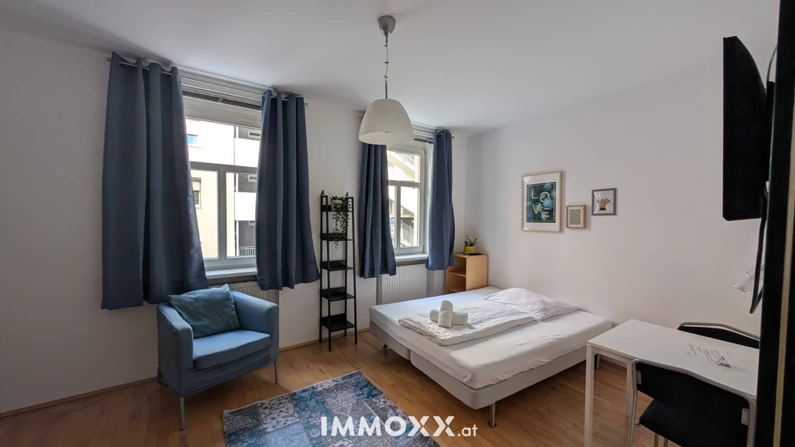 Gemütliches Studio-Apartment mit Bett, Sessel und zwei Fenstern für viel Tageslicht.