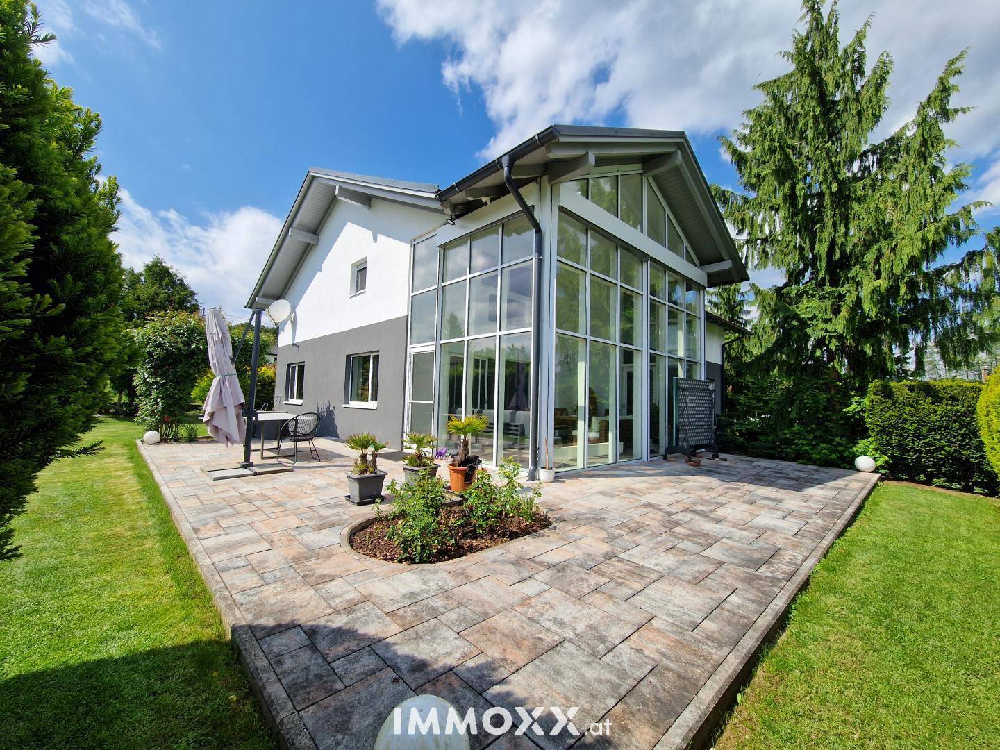 Moderne Architektur des Hauses mit großer Glasfassade, gepflasterter Terrasse und weitläufigem Garten.