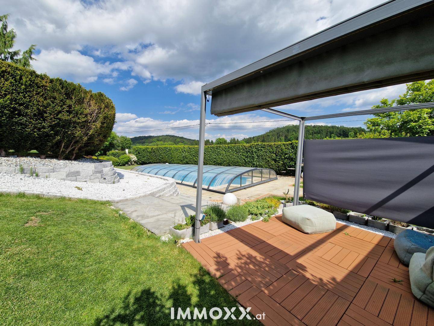 Gemütlicher Sitzbereich im Freien mit Holzfliesen und Pergola, Blick auf den Pool und die Landschaft.