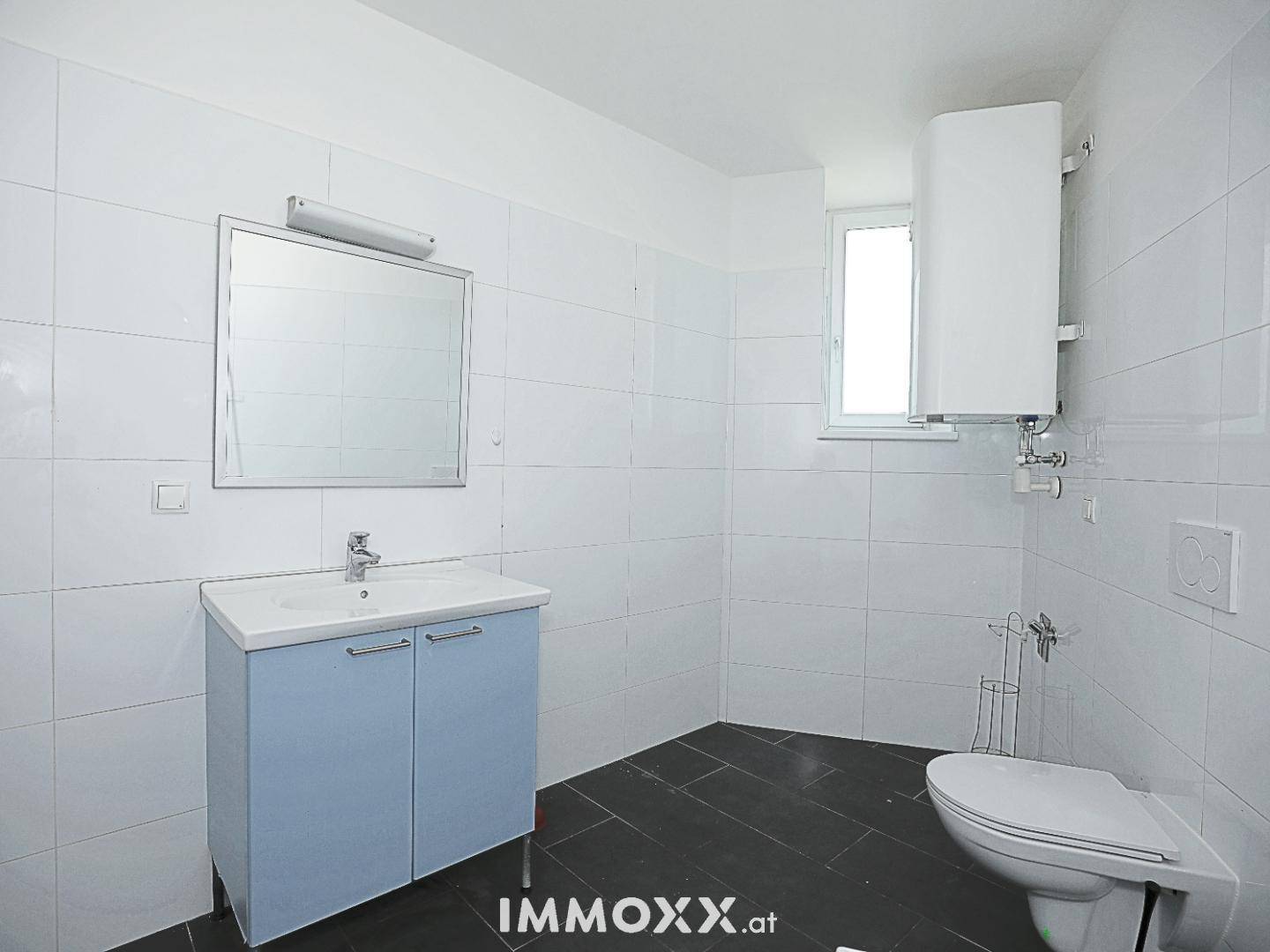 Badezimmer mit Waschbecken, Toilette, Fenster und einem Warmwasserboiler an der Wand.