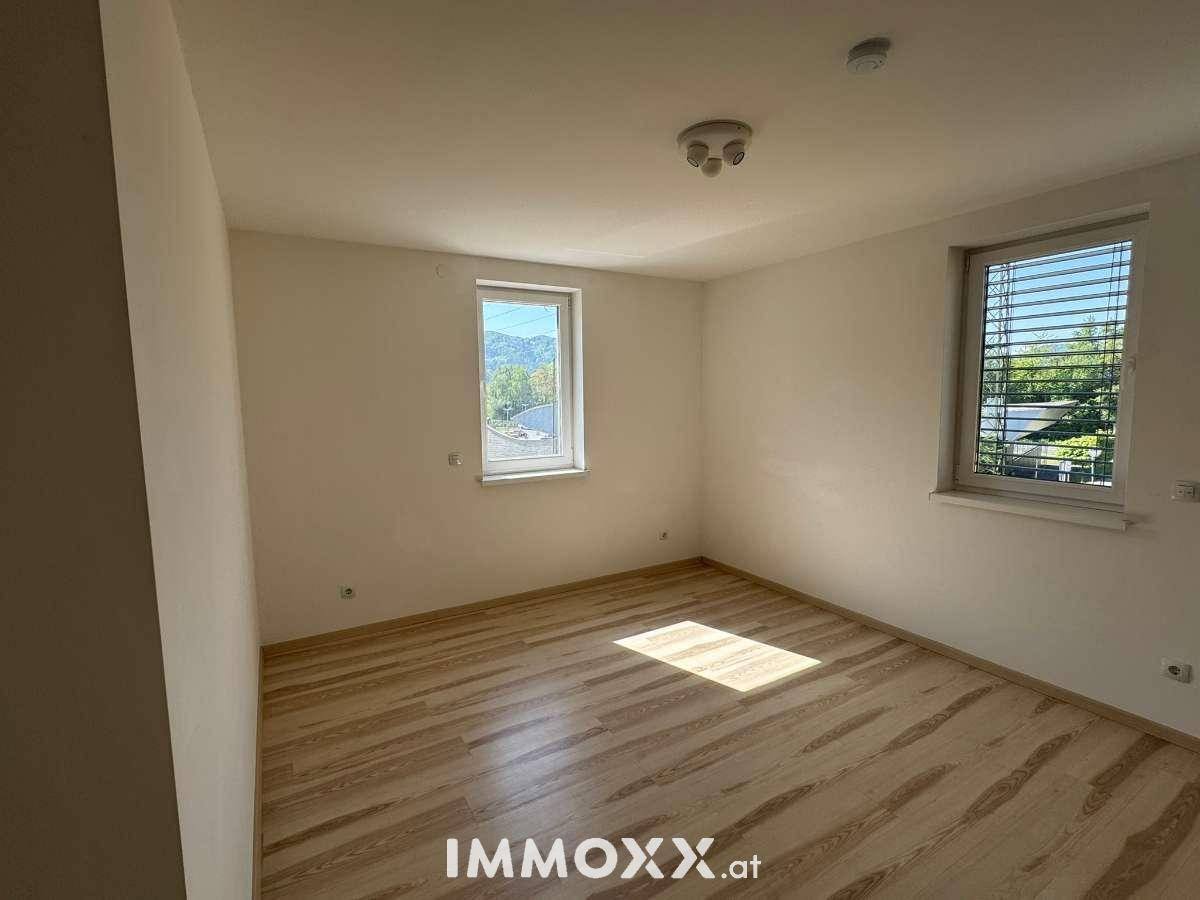 Helles, leeres Zimmer mit Laminatboden und zwei Fenstern, eines davon mit Bergblick.