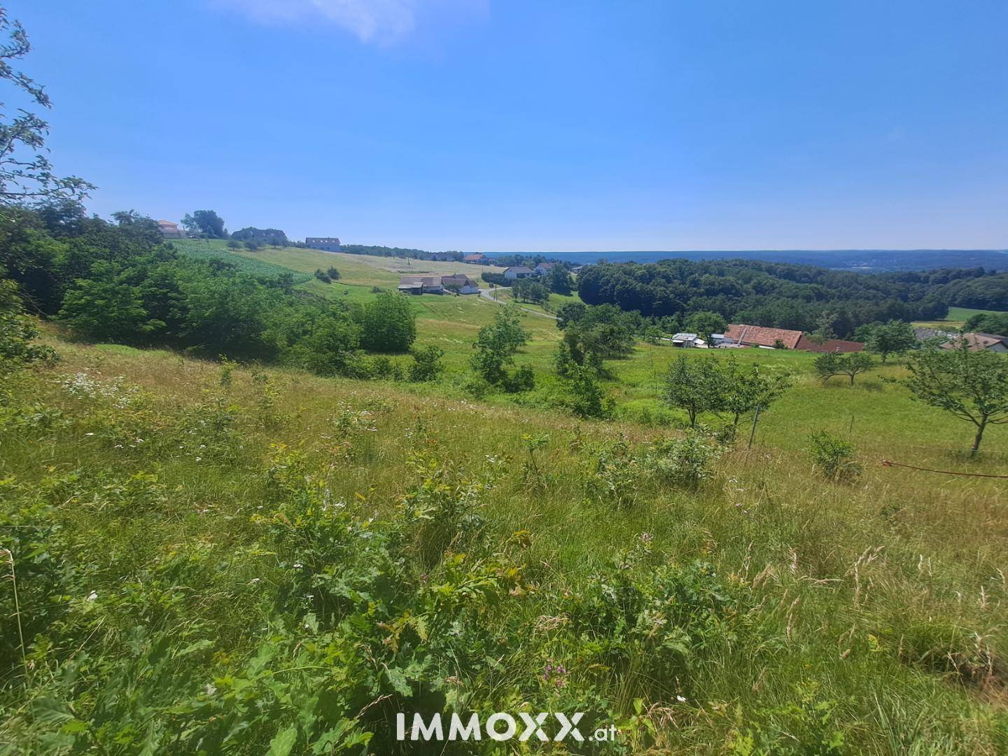 Weitläufige grüne Landschaft mit sanften Hügeln und vereinzelten Häusern unter blauem Himmel.