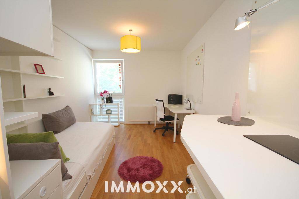 Helles Studio-Apartment mit Bett, Schreibtisch und einem Fenster, das viel Tageslicht hereinlässt.