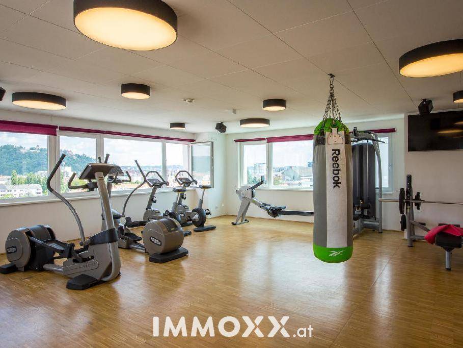 Gut ausgestatteter Fitnessraum mit verschiedenen Trainingsgeräten und großen Fenstern für Tageslicht.