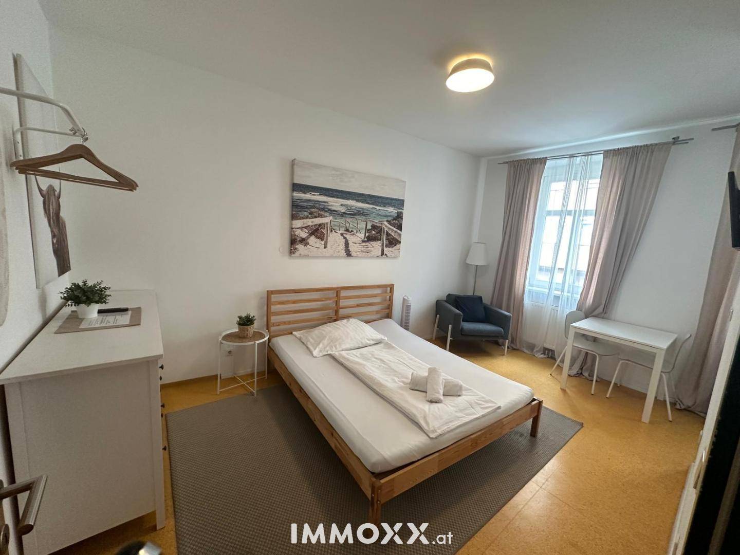 Gemütliches Schlafzimmer mit großem Fenster, Vorhängen und einem kleinen Arbeitsbereich.