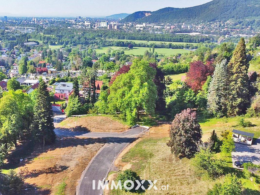 Luftaufnahme einer grünen Hügellandschaft mit neuer Straße, Bäumen und Fernblick auf eine Stadt und Berge.