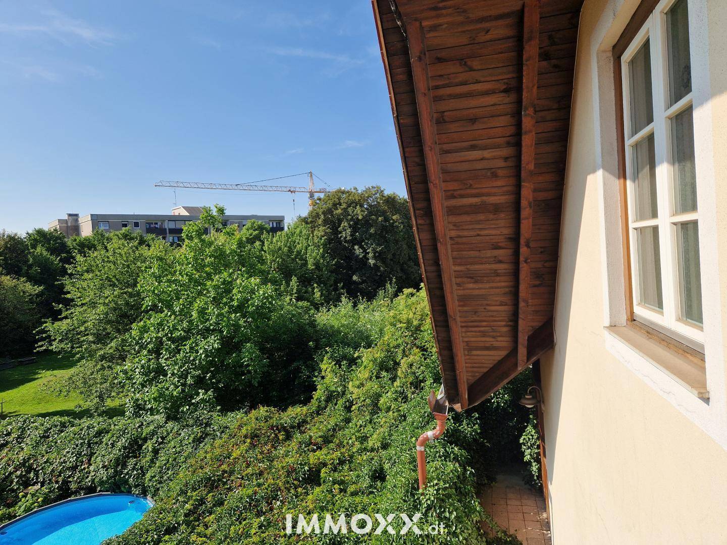 Blick aus dem Fenster auf einen kleinen blauen Pool, umgeben von Bäumen und einem Kran in der Ferne.