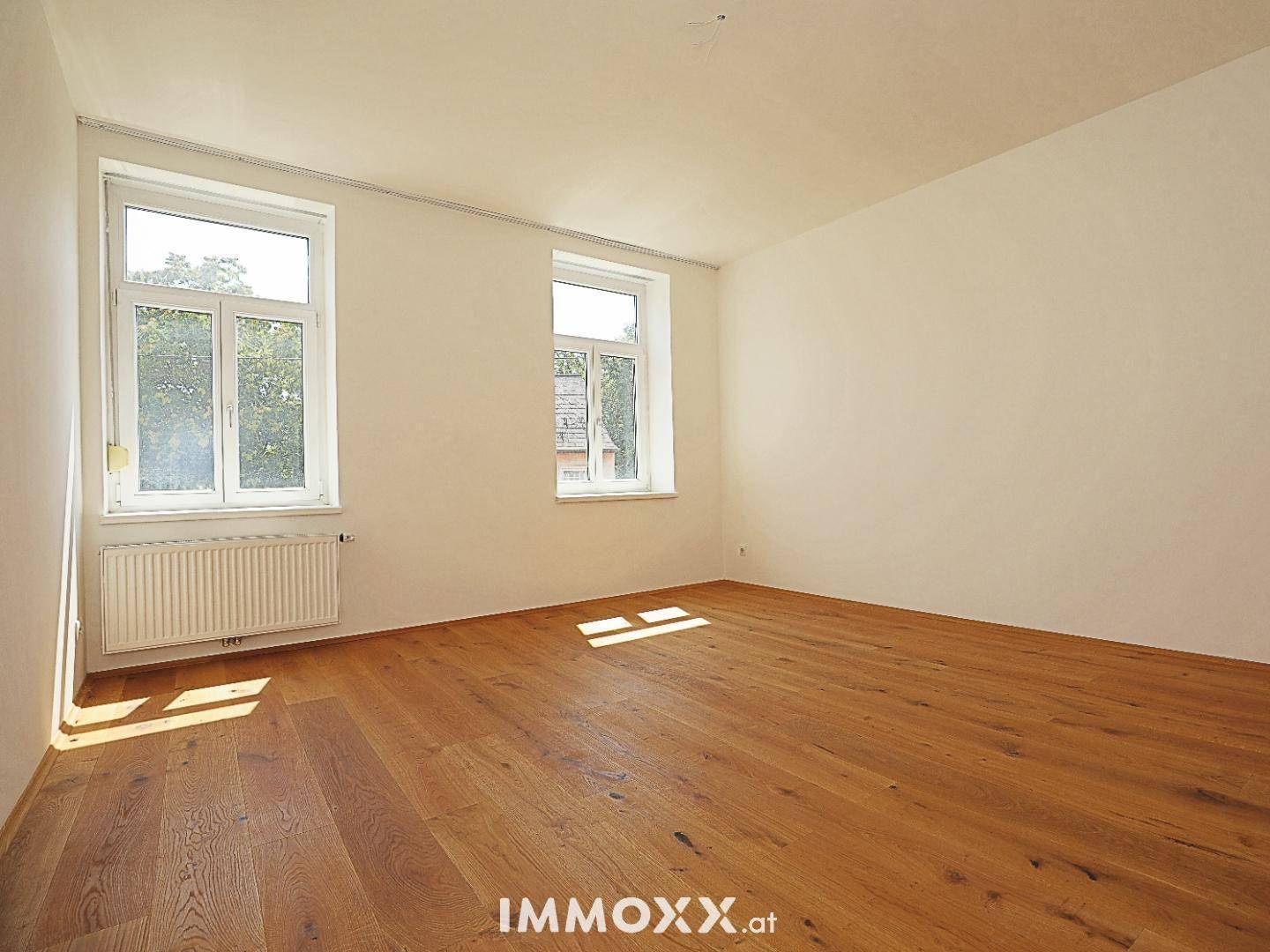 Heller, leerer Raum mit Holzboden und zwei Fenstern, die viel Tageslicht hereinlassen.