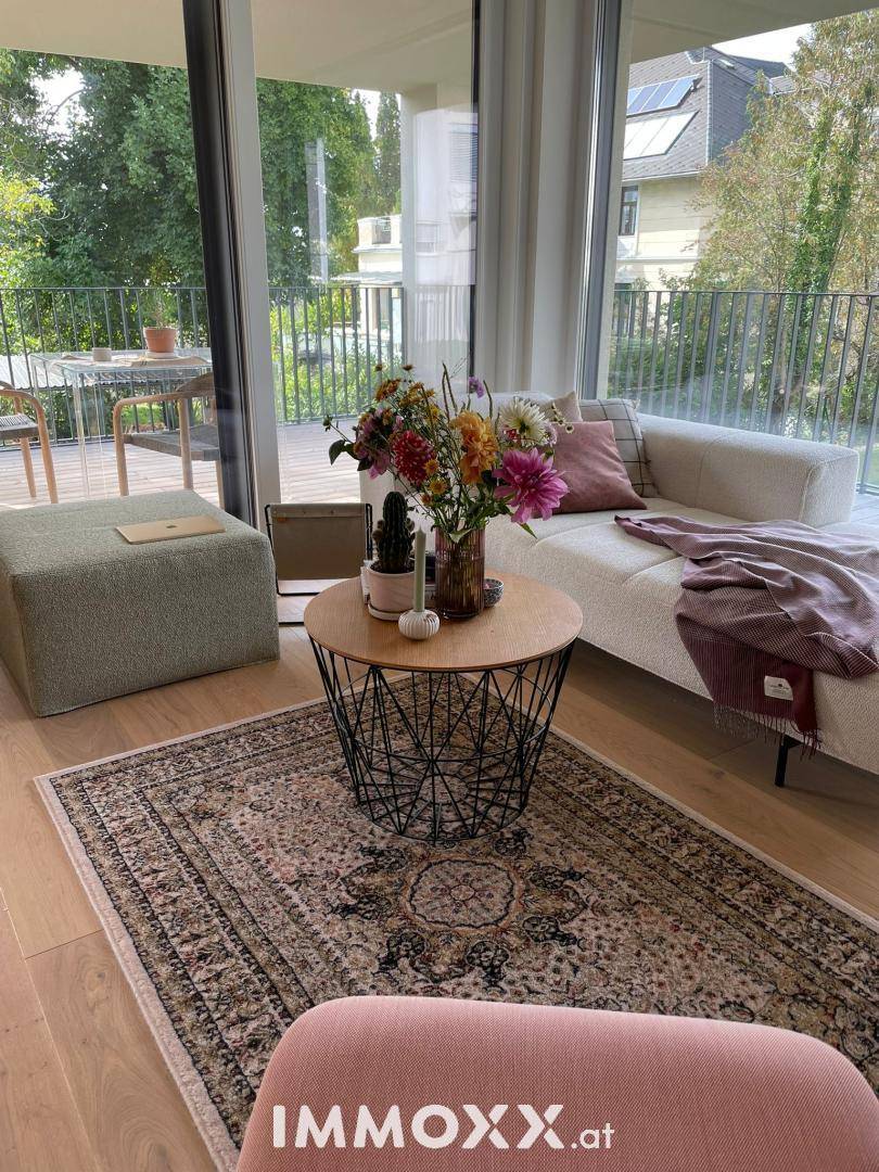 Gemütlicher Wohnbereich mit hellem Sofa, Teppich und großen Fenstern mit Blick auf den Balkon und Bäume.