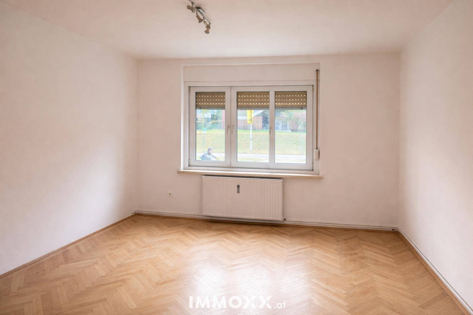Leerer Raum mit Fischgrätparkett, weißen Wänden und einem Fenster mit Rollladen.