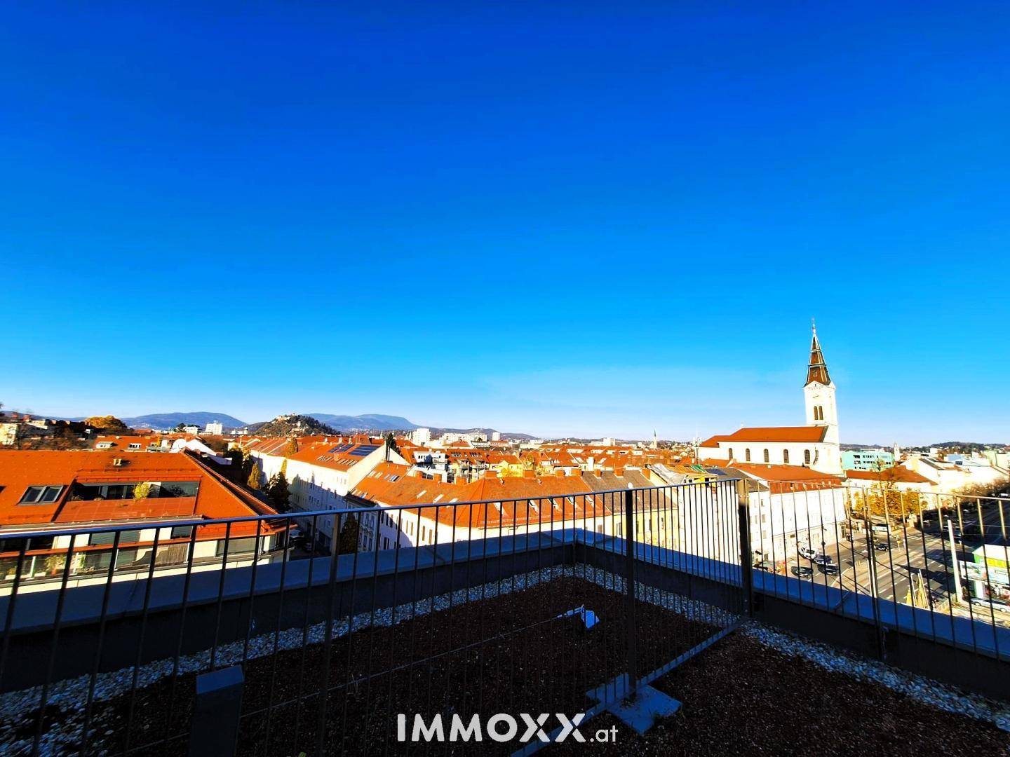 Weitläufiger Blick über die Stadt mit Kirchturm und Bergen im Hintergrund unter klarem Himmel.