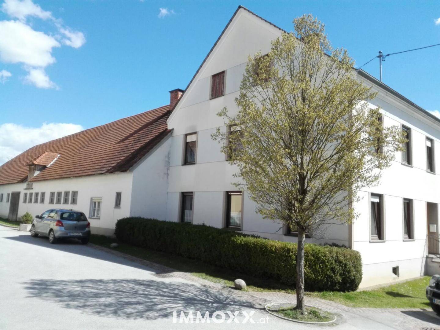 Weißes Wohnhaus mit braunem Ziegeldach und einem Baum im Vorgarten unter blauem Himmel.