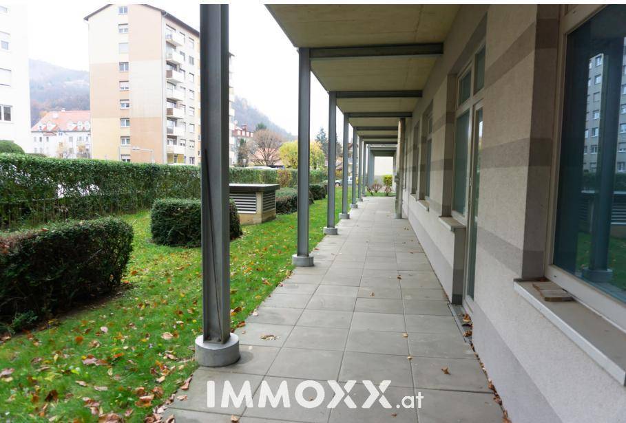 Außenansicht eines Gebäudes mit überdachtem Gehweg, Rasenfläche und gepflegten Hecken.
