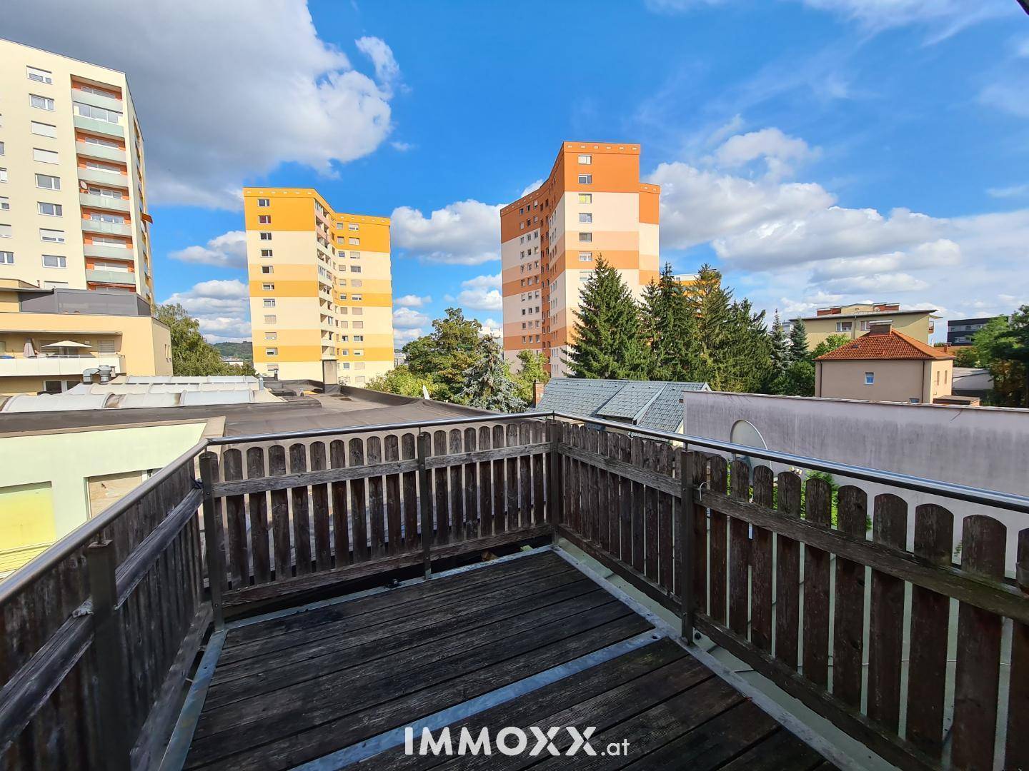 Hölzerne Dachterrasse mit Geländer und weitem Blick auf umliegende Gebäude und Bäume.