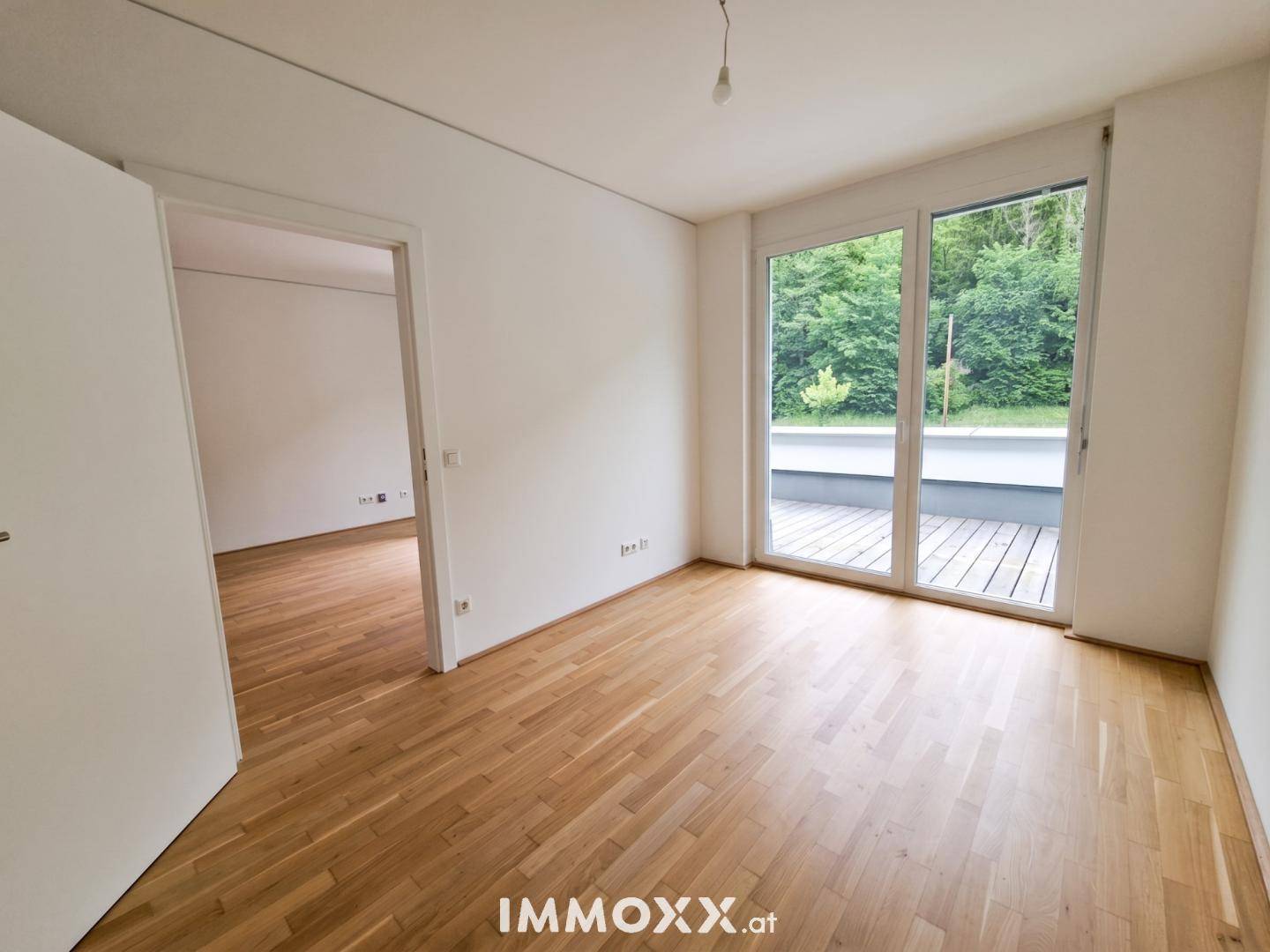 Leerer Raum mit Parkettboden, großen Fenstern und Zugang zu einem Außenbereich, mit offener Tür zu einem weiteren Zimmer.
