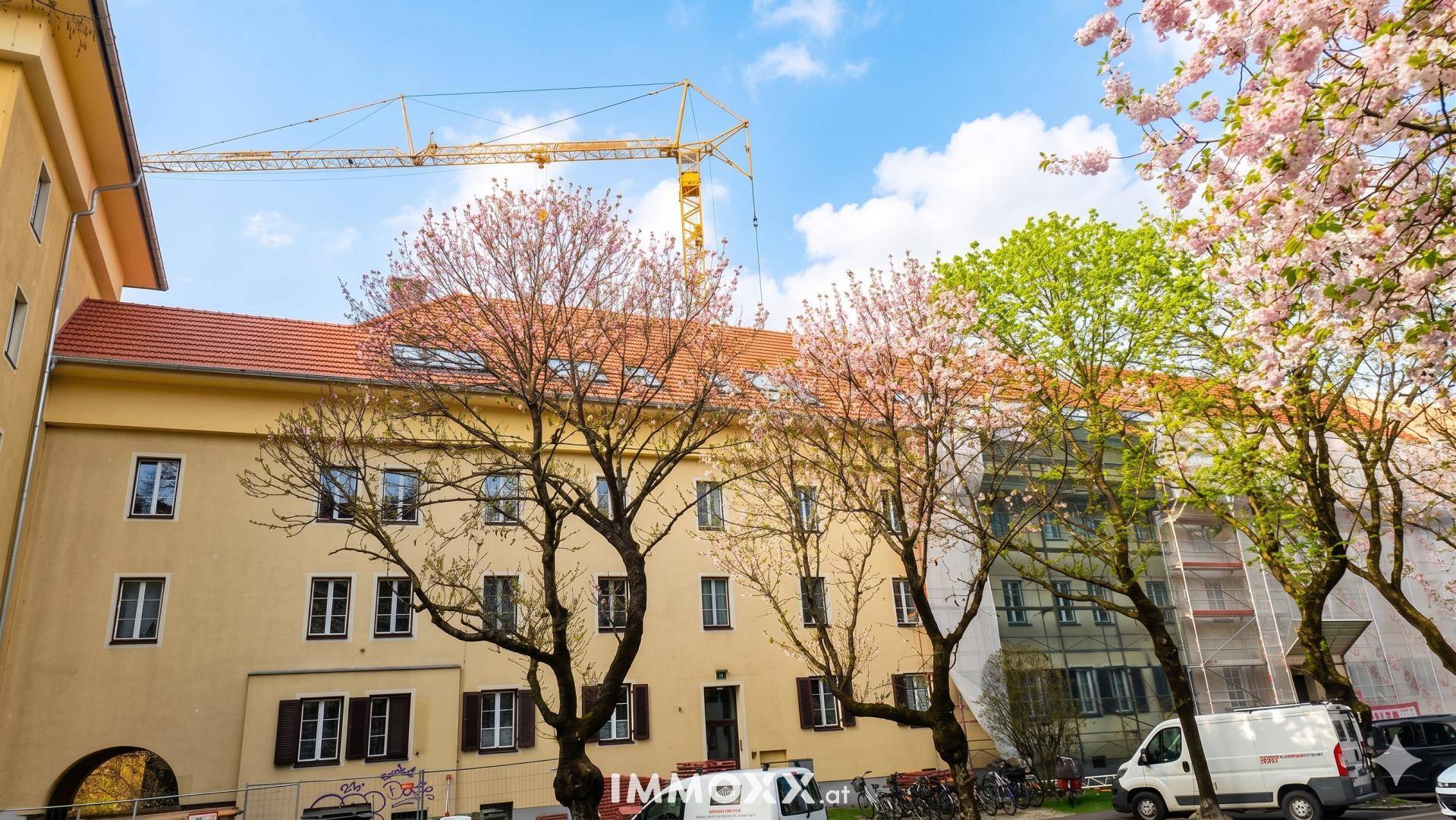 Gebäudefassade mit blühenden Bäumen im Vordergrund und einem Baukran im Hintergrund.