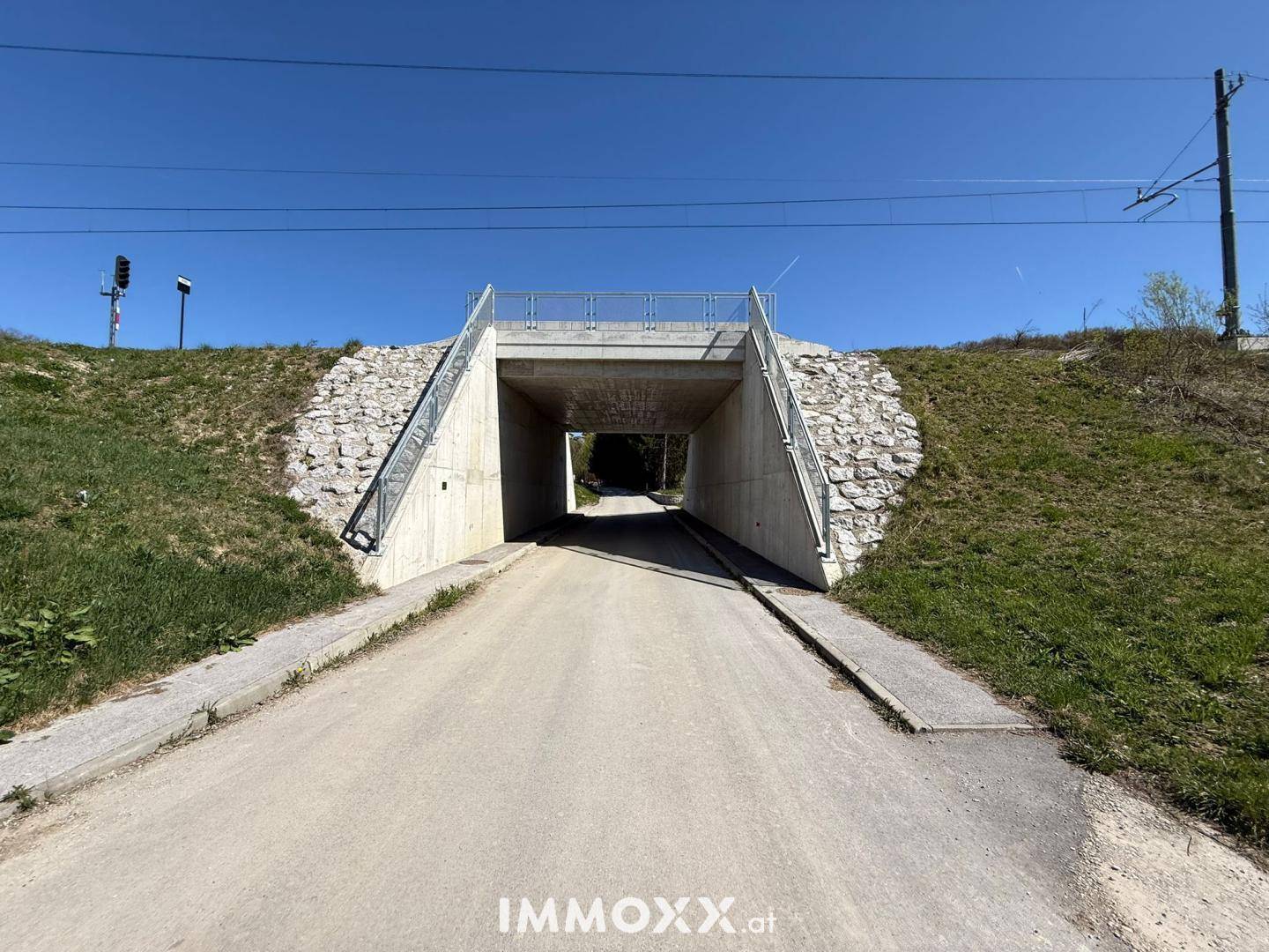Eine moderne Unterführung aus Beton mit Treppen und einem Schotterweg, umgeben von grünen Böschungen.