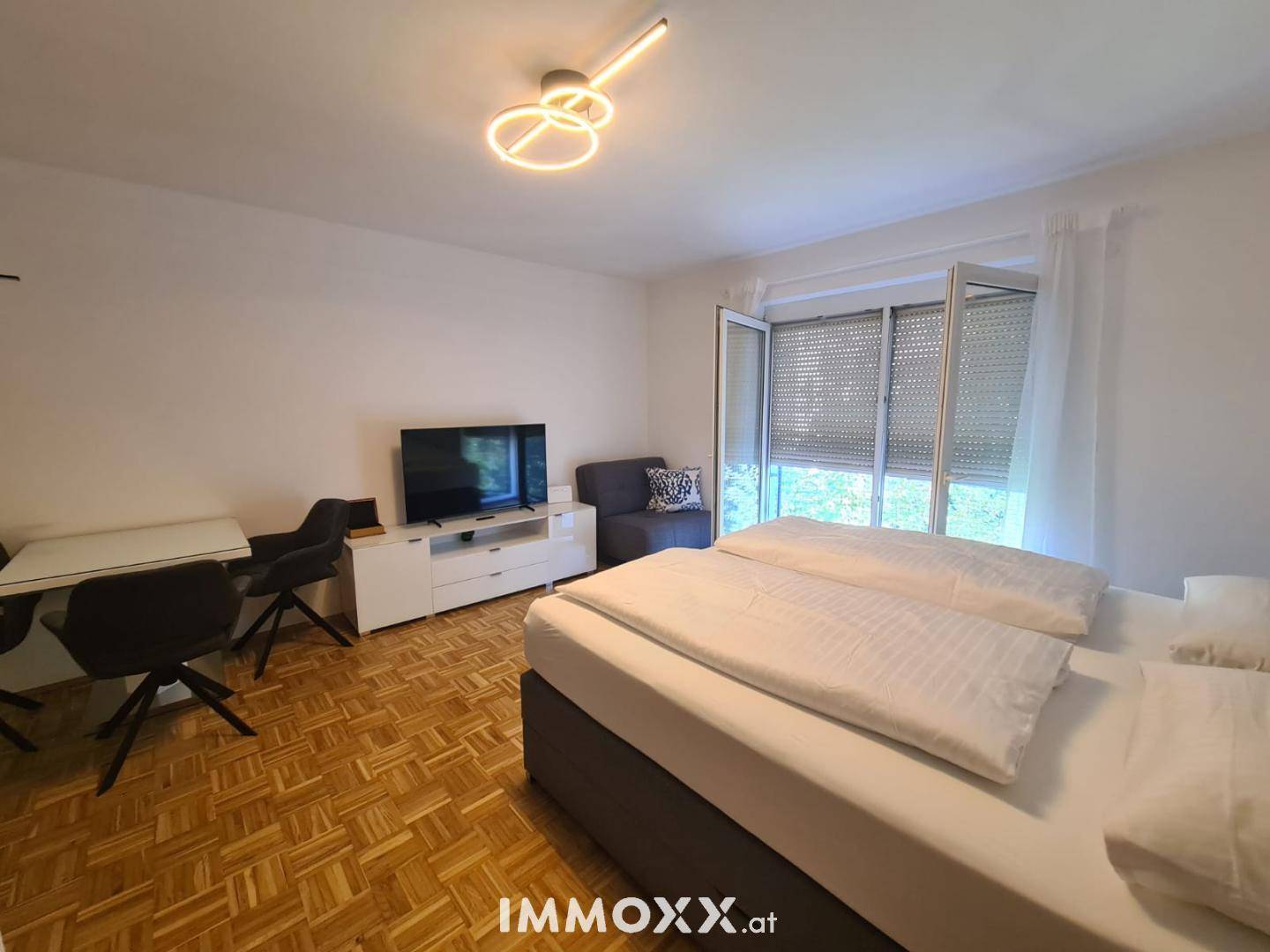 Geräumiges Studio-Apartment mit Schlafbereich, Wohnbereich und Blick ins Grüne durch große Fenster.